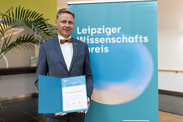 Prof. Alex Stahr Mann mit Urkunde steht vor Plakat