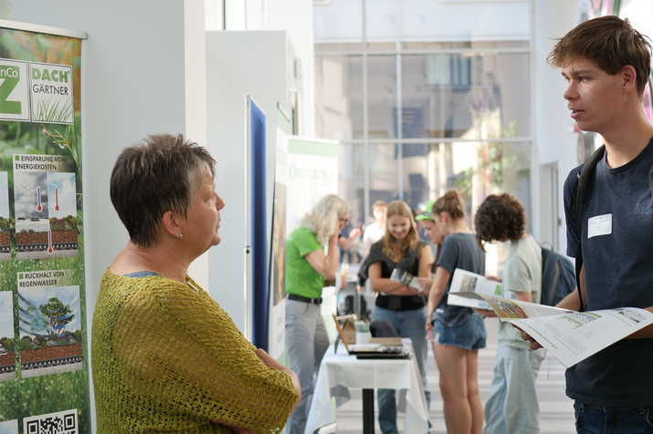 Schüler und Mitarbeiterin am Infostand unterhalten sich, Schüler hat Broschüre in der Hand